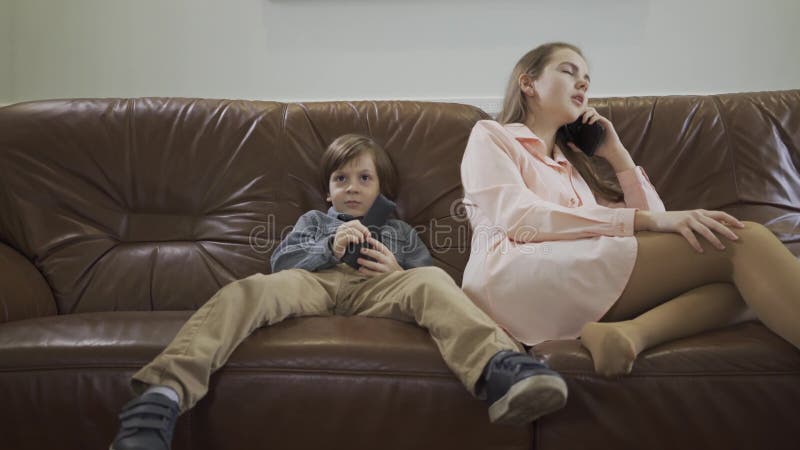 Il piccolo ragazzo sveglio che si siede sul sofà di cuoio con le gambe a parte che cambiano i canali sulla TV facendo uso della s