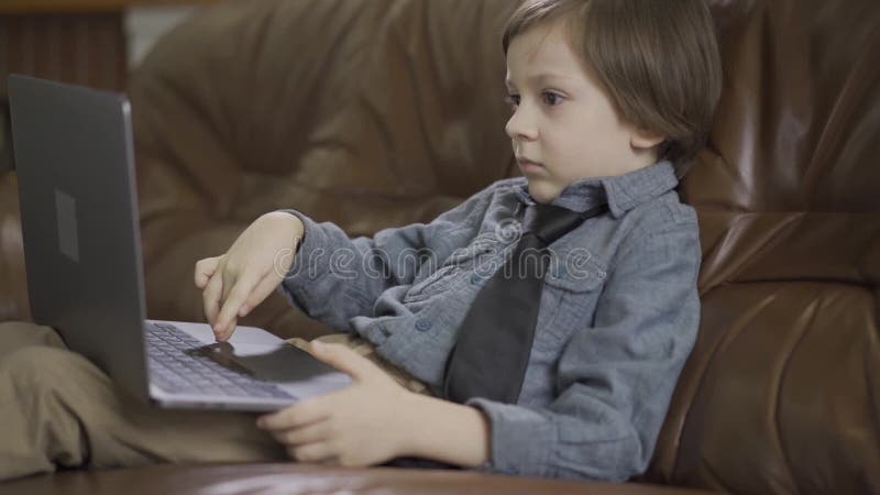 Il ragazzino in rivestimento dei jeans che si siede sul sof? di cuoio che gioca sul computer portatile Lo svago del bambino moder
