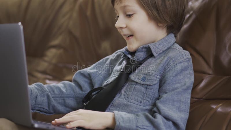 Il ragazzino in rivestimento dei jeans che si siede sul sof? di cuoio che gioca sul computer portatile che sorride felicemente Lo
