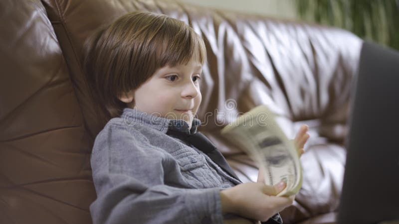 Il ragazzino in rivestimento dei jeans che si siede sul sofà di cuoio davanti al computer portatile che gioca con i soldi Un pacc