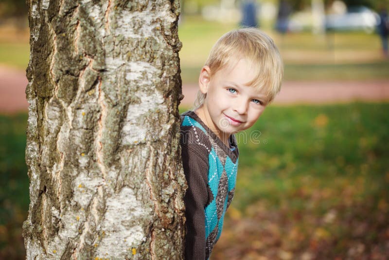 Il ragazzino sveglio del ritratto in un maglione tricottato sta giocando dietro un albero nel parco di autunno, gioco al nascondi