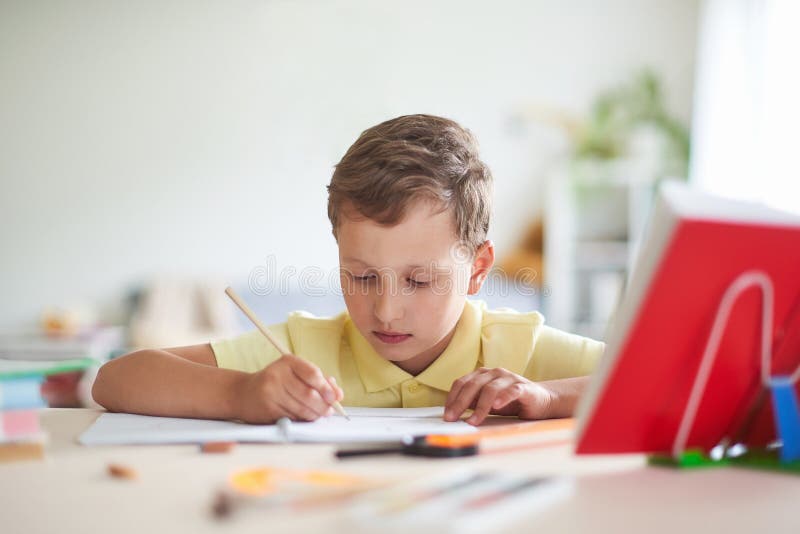 Il ragazzo fa il suo compito a casa il bambino felice alla tavola con i rifornimenti di scuola ha concentrato la scrittura nella immagine stock Il ragazzo fa il suo compito a casa il bambino felice alla tavola con i rifornimenti di scuola ha concentrato la scrittura nella immagine stock
