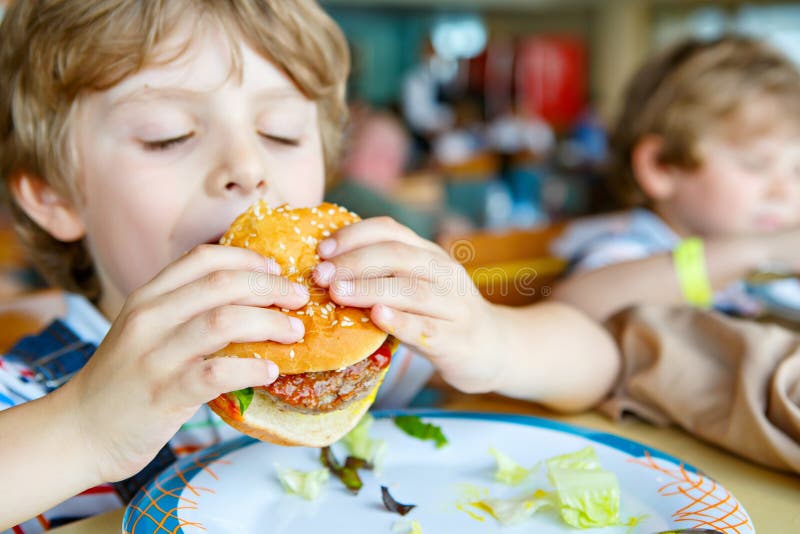 Il ragazzo prescolare in buona salute sveglio del bambino mangia l'hamburger che si siede in caffè della scuola materna o della s immagine stock libera da diritti Il ragazzo prescolare in buona salute sveglio del bambino mangia l'hamburger che si siede in caffè della scuola materna o della s immagine stock libera da diritti