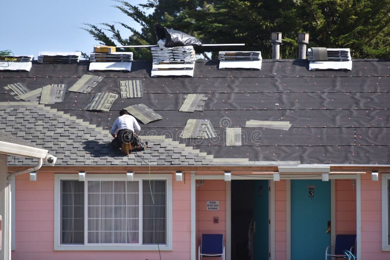 Male roofer stapling asphalt roof tiles to repair a roof on a motel. Motel is pink in color with blue doors and white trim. Roof tiles are grey in color. Male roofer stapling asphalt roof tiles to repair a roof on a motel. Motel is pink in color with blue doors and white trim. Roof tiles are grey in color.