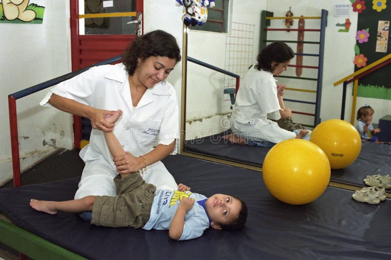 Brazil, Pernambuco, city Gravata: a woman doctor is to stimulate motor skills of a child who has physical and mental disabilities. The physio is stretching a leg of the boy. They are working, training, in a therapy room with toys. Mirror image of these people in the big mirror on the wall. Brazil, Pernambuco, city Gravata: a woman doctor is to stimulate motor skills of a child who has physical and mental disabilities. The physio is stretching a leg of the boy. They are working, training, in a therapy room with toys. Mirror image of these people in the big mirror on the wall.