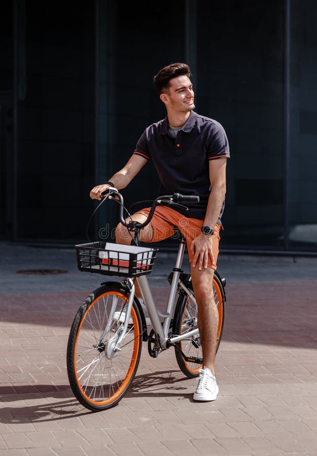 Il tipo alla moda vestito nei giri d'avanguardia dell'abbigliamento casual va in bicicletta nella via contro lo sfondo di una cos fotografia stock libera da diritti Il tipo alla moda vestito nei giri d'avanguardia dell'abbigliamento casual va in bicicletta nella via contro lo sfondo di una cos fotografia stock libera da diritti