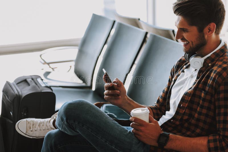 Il tipo allegro sta godendo del telefono e del caffè all'aeroporto fotografia stock Il tipo allegro sta godendo del telefono e del caffè all'aeroporto fotografia stock