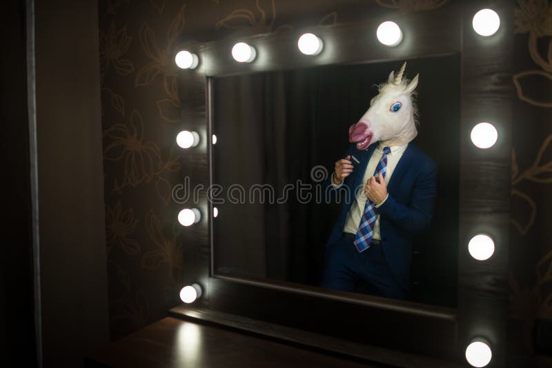 Glamour guy in elegant suit and comical mask looks at himself in the mirror in dressing room. Freaky young man in stylish room posing like a boss. Unusual unicorn. Glamour guy in elegant suit and comical mask looks at himself in the mirror in dressing room. Freaky young man in stylish room posing like a boss. Unusual unicorn.