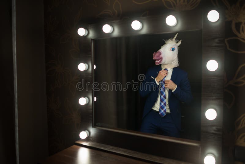 Glamour guy in elegant suit and comical mask looks at himself in the mirror in dressing room. Freaky young man in stylish room posing like a big boss. Unusual unicorn. Glamour guy in elegant suit and comical mask looks at himself in the mirror in dressing room. Freaky young man in stylish room posing like a big boss. Unusual unicorn.