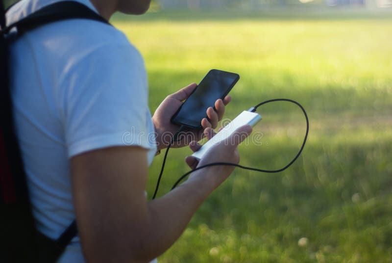 Il tipo sta tenendo un caricatore portatile con uno smartphone in sua mano Uomo su un fondo della natura con i verdi immagine stock Il tipo sta tenendo un caricatore portatile con uno smartphone in sua mano Uomo su un fondo della natura con i verdi immagine stock