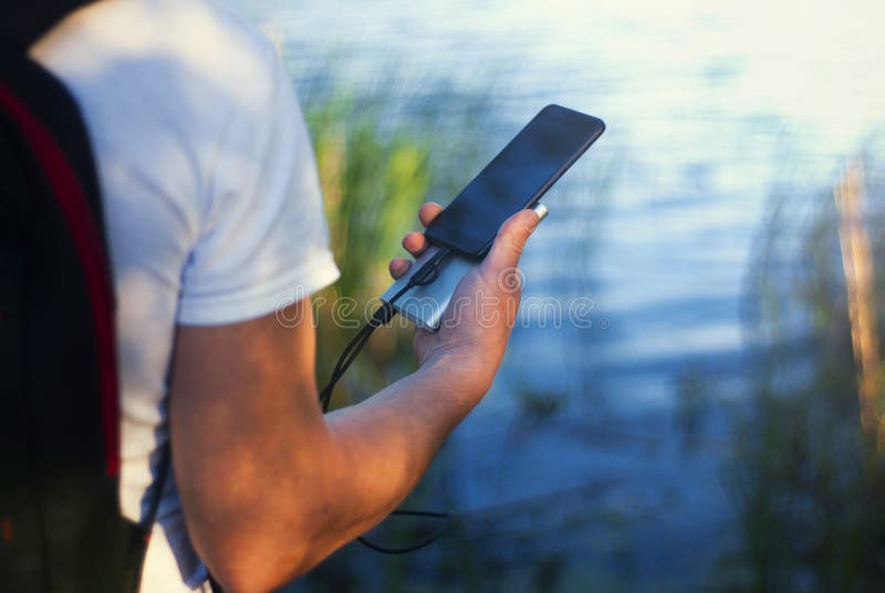 Il tipo sta tenendo un caricatore portatile con uno smartphone in sua mano Uomo su un fondo della natura con una pianta e un lago fotografia stock libera da diritti Il tipo sta tenendo un caricatore portatile con uno smartphone in sua mano Uomo su un fondo della natura con una pianta e un lago fotografia stock libera da diritti
