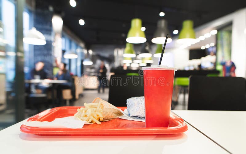 Red tray with Fast Food menu is on the table in the restaurant. French fries, burger, red glass with a cola in the tray against the background of a fast food restaurant. Harmful, caloric food. Red tray with Fast Food menu is on the table in the restaurant. French fries, burger, red glass with a cola in the tray against the background of a fast food restaurant. Harmful, caloric food