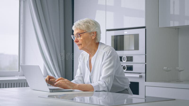 Il vecchio manager della società sceglie il laptop grigio che si trova su un grande tavolo bianco in cucina contro le finestre con fotografie stock