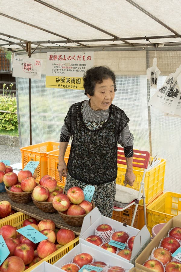 Takayama, Japan - September 24, 2016: Older active lady who is a vendor on the public morning market in front of the Jinya Hall promotes her red apples. Black clothes, white tarp background. Takayama, Japan - September 24, 2016: Older active lady who is a vendor on the public morning market in front of the Jinya Hall promotes her red apples. Black clothes, white tarp background.