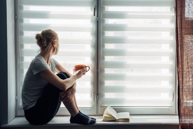 Illness young girl on home quarantine. Woman sits on windowsill, reads a book, drinks tea. Coronavirus pandemic, epidemic in