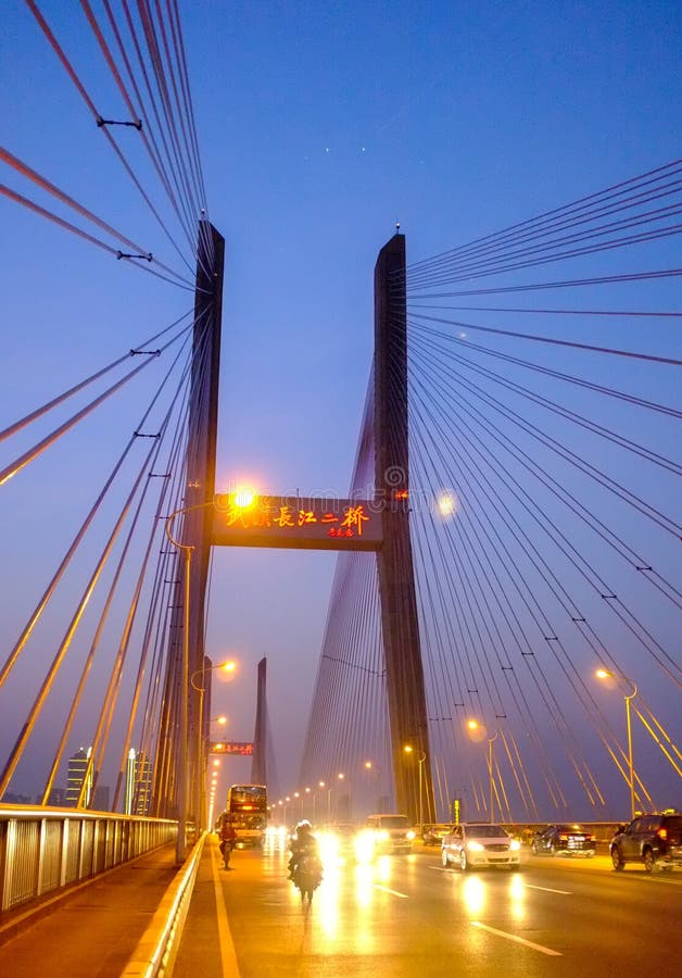 A cable-stayed bridge at twilight features two illuminated pylons with radiating support cables and warm streetlights lining the roadway, busy with cars, motorcycles, and a truck moving in both directions beneath a red-lit digital signboard displaying Chinese characters. Ideal for infrastructure editorials, urban architecture portfolios, transportation articles, and cityscape headers, shot to emphasize structural geometry, evening lighting, and dynamic traffic activity. Illuminated pylons stock images, royalty-free photos and pictures