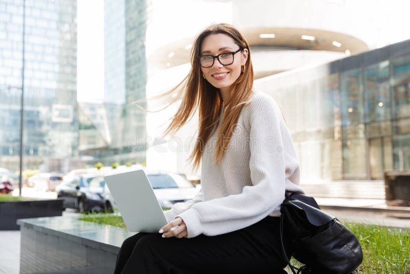 Beautiful young happy business woman posing sitting outdoors near business center wearing eyeglasses using laptop computer