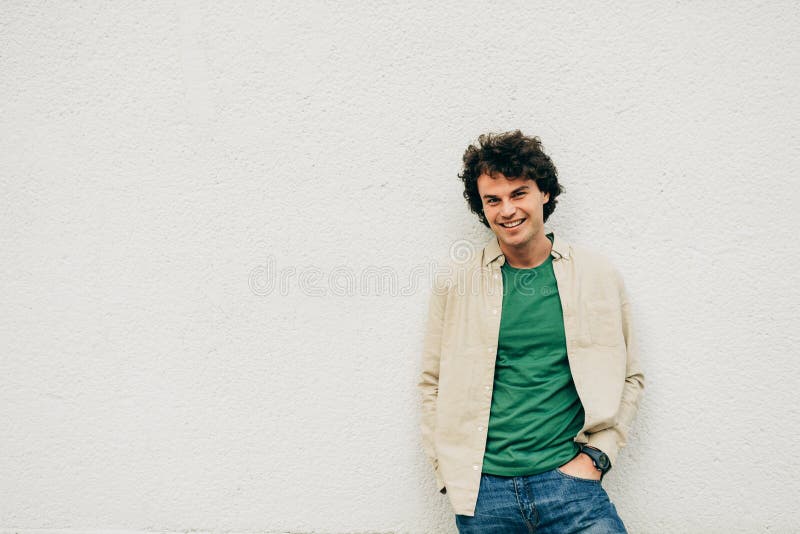 Image of cheerful young man posing for advertisement, smiling and looking at the camera, standing at building concrete wall