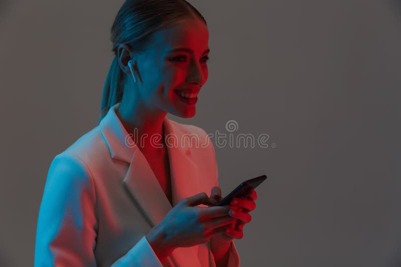 Image closeup of attractive woman 20s wearing earpods holding and using mobile phone while standing under neon lights