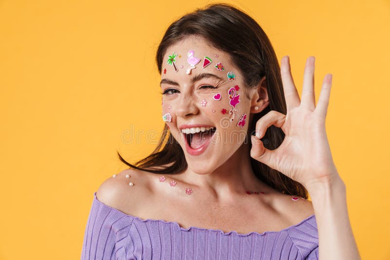 Image d'une femme heureuse gestant ok signe et winking. Image d'une femme heureuse avec des autocollants sur le visage gestant ok signe et se réveillant isolé sur le mur jaune