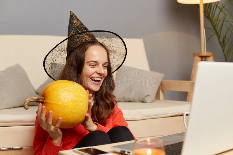 Image of excited happy optimistic young adult woman in witch hat having online video call on laptop, celebrating halloween at home, showing pumpkin to notebook display. Image of excited happy optimistic young adult woman in witch hat having online video call on laptop, celebrating halloween at home, showing pumpkin to notebook display