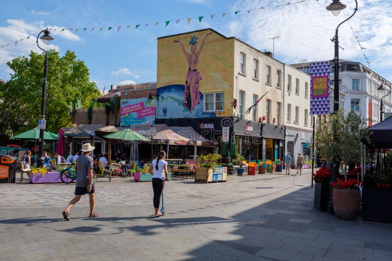 The image features a lively square with outdoor seating, colourful signage, and a mural of a dancer on a building. People are walking and using scooters, enjoying a sunny day. Brightly coloured banners and bunting add to the festive atmosphere. A restaurant with outdoor tables and umbrellas is visible, surrounded by potted plants and hanging lights. The architecture reflects a classic urban style, contributing to the dynamic and bustling environment. Dynamic colour stock images, royalty-free photos and pictures