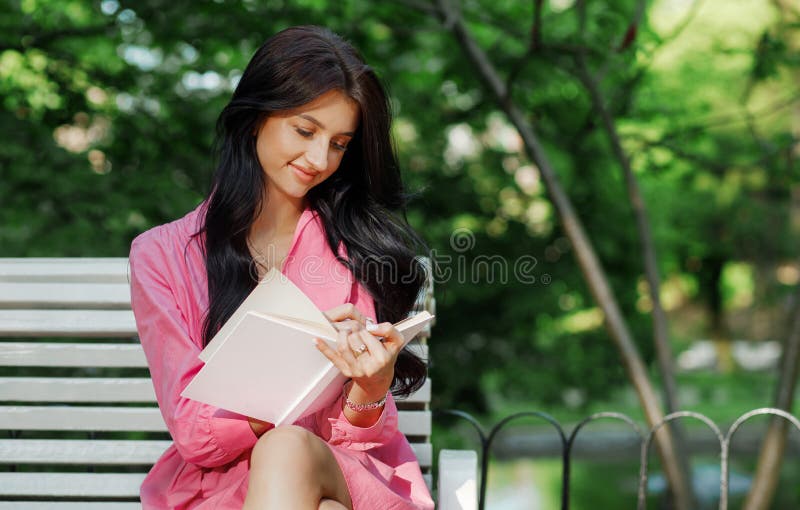 Image of a happy smiling young female student sitting outdoors in a nature park with a book in her hands.