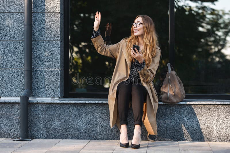 Image of joyous woman using mobile phone while sitting in front of building. Image of joyous woman 20s wearing coat and eyeglasses using mobile phone while royalty free stock photo Image of joyous woman using mobile phone while sitting in front of building. Image of joyous woman 20s wearing coat and eyeglasses using mobile phone while royalty free stock photo