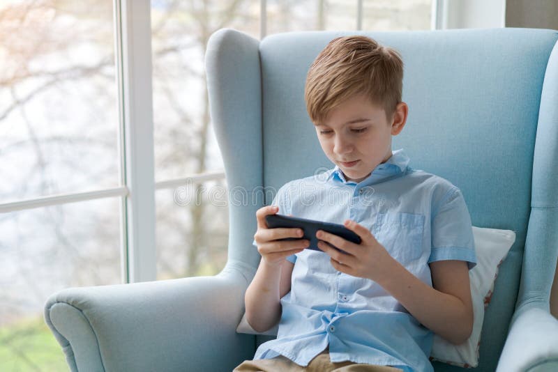 Image of little cheerful boy holding smartphone and playing game while sitting