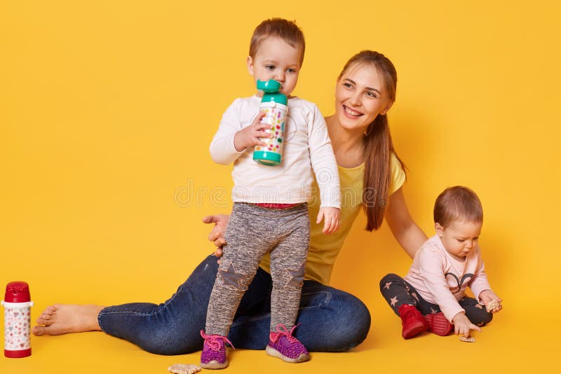 Image of loving attractive mother looks after her little children, twins playing with mommy. Playful kids drink tasty baverage stock photography Image of loving attractive mother looks after her little children, twins playing with mommy. Playful kids drink tasty baverage stock photography