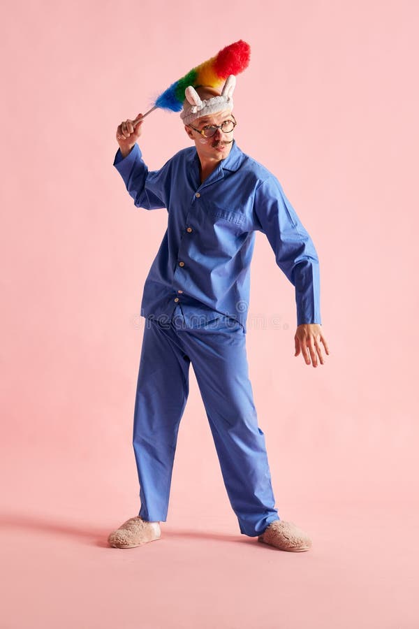 Image of man in ridiculous headband and nightwear holding dust brush and grimacing over pastel rose background.