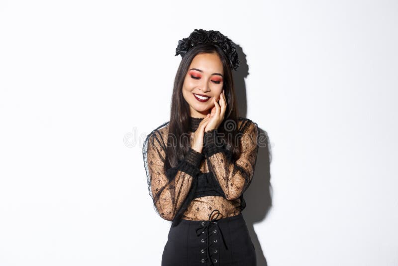 Image of sensual, beautiful asian woman in gothic lace dress and black wreath smiling coquettish, touching face and