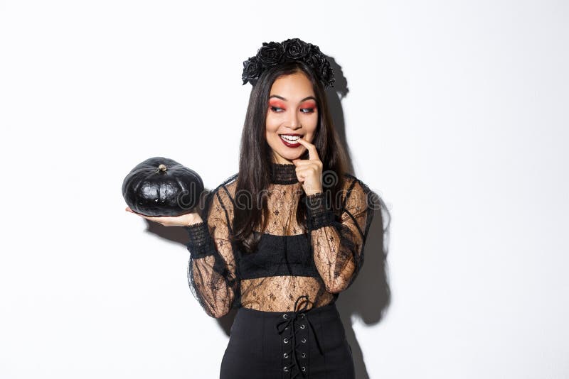 Image of sexy asian woman in witch costume looking at black pumpking and smiling, standing over white background