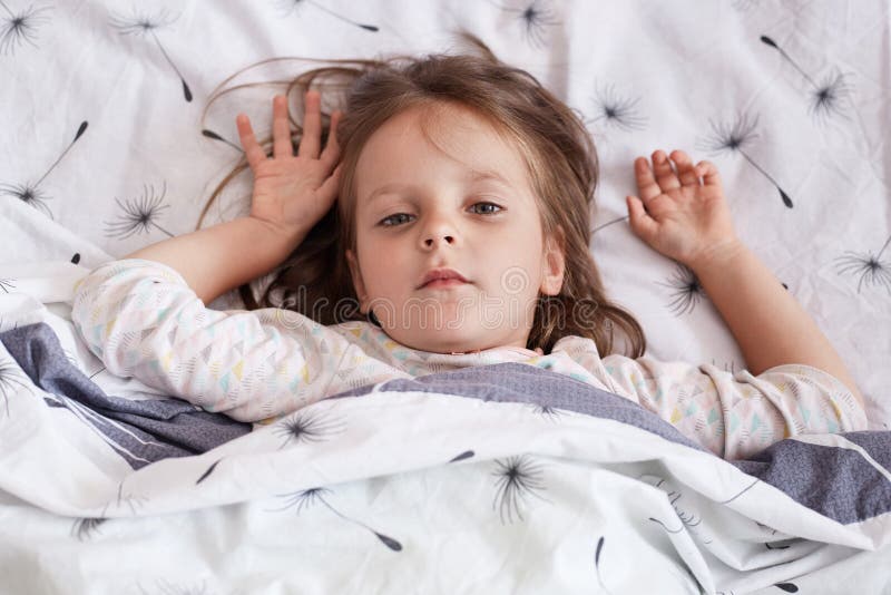 Image of sleepy sweet beautiful little girl lying in her bed, raising her arms, looking directly at camera, having serious facial