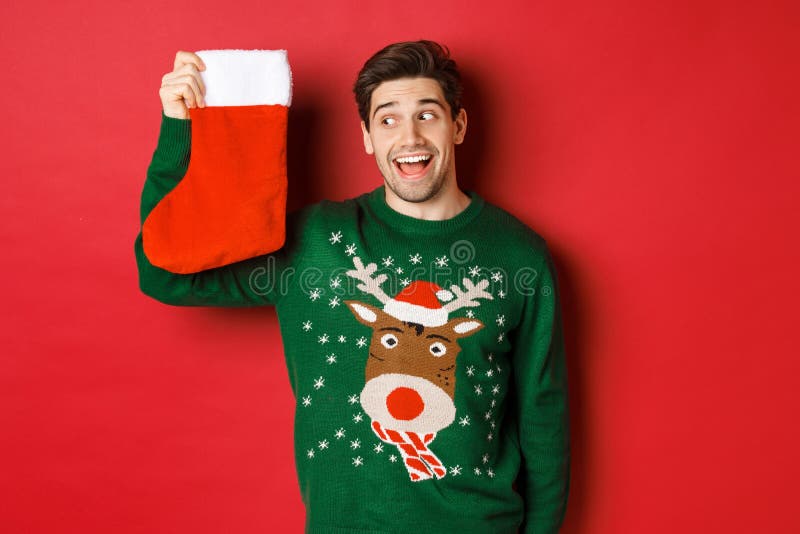 Image of surprised and amused man in green sweater, looking at christmas stocking with presents and smiling, standing