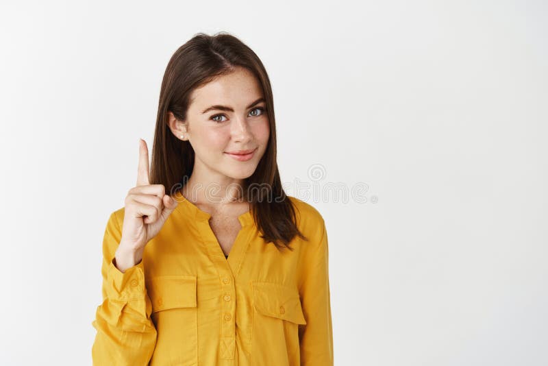 Image of young thoughtful woman showing advertisement, pointing finger up and smiling at camera, giving knowing look