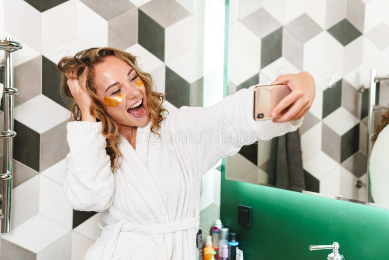 Image of young beautiful blond woman in white housecoat with eye patches taking selfie photo in bathroom. Image of young beautiful blond woman in white housecoat with eye patches taking selfie photo in bathroom