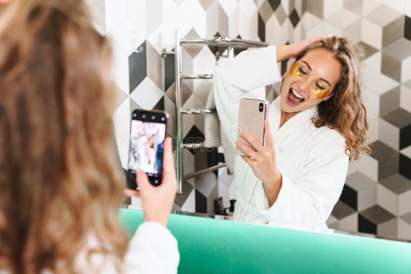 Image of young beautiful blond woman in white housecoat with eye patches taking selfie photo in bathroom. Image of young beautiful blond woman in white housecoat with eye patches taking selfie photo in bathroom