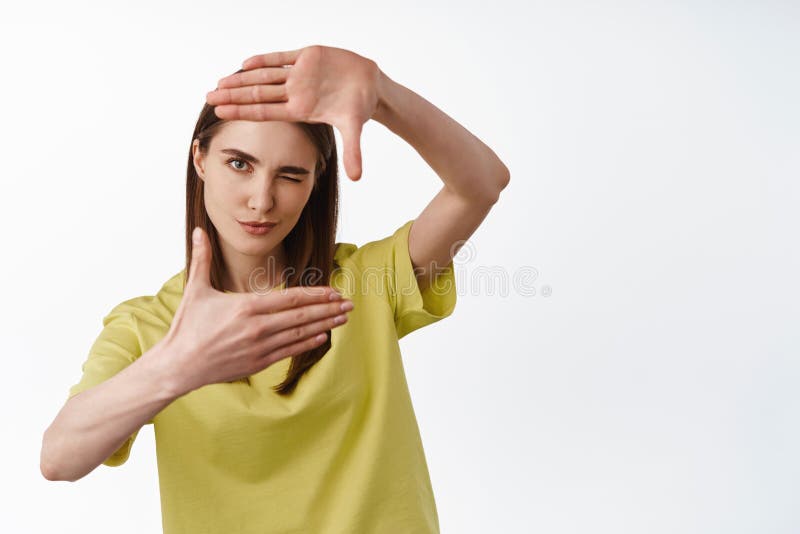 Image of young woman looks through frame hands, measure smth, thinking of perfect angle, camera shot, searching for inspiration, stand against white background. Image of young woman looks through frame hands, measure smth, thinking of perfect angle, camera shot, searching for inspiration, stand against white background.