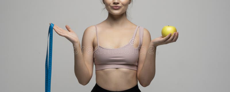 Crop image of attractive woman in a sport wear is smile and looks in a camera while holds blue measuring tape and an apple. Health care and healthy nutrition. Perfect slim body. Crop image of attractive woman in a sport wear is smile and looks in a camera while holds blue measuring tape and an apple. Health care and healthy nutrition. Perfect slim body