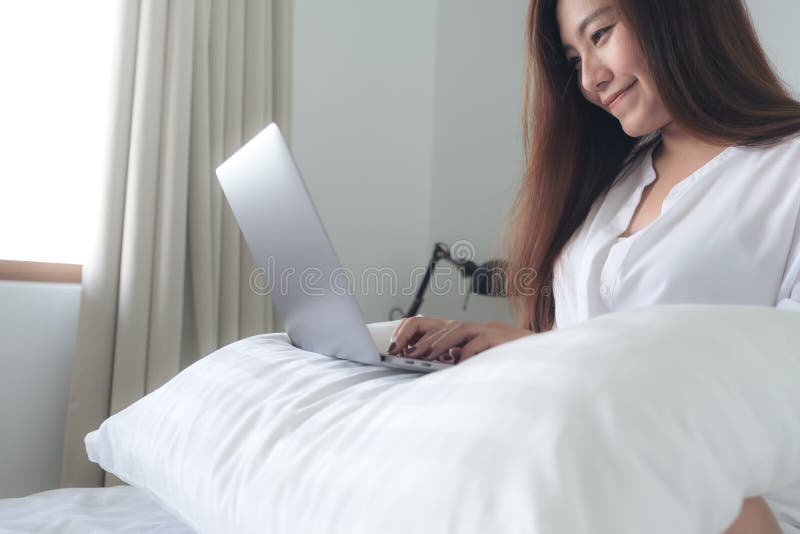 Imagem do close up de uma mulher asiática bonita que senta-se em uma cama, usando-se e datilografando no portátil fotografia de stock royalty free Imagem do close up de uma mulher asiática bonita que senta-se em uma cama, usando-se e datilografando no portátil fotografia de stock royalty free