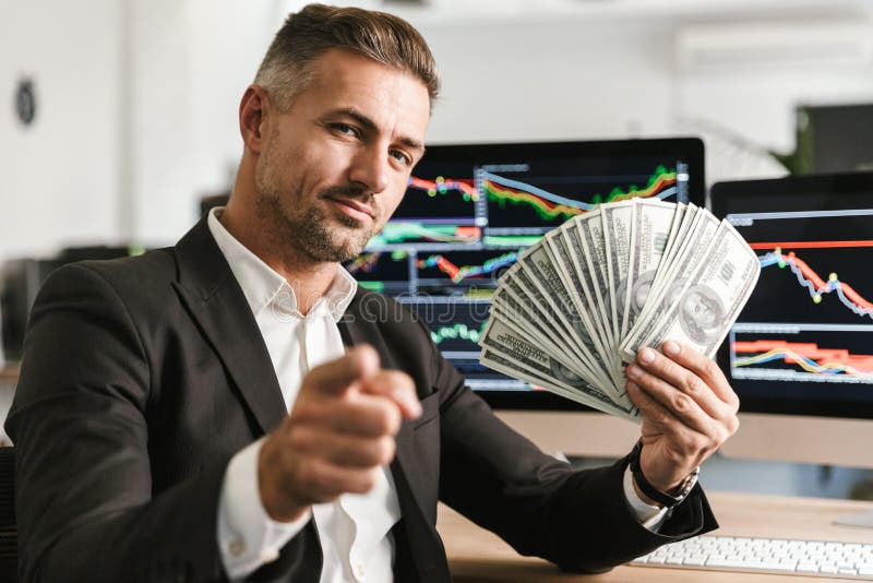 Image of pleased businessman 30s wearing suit holding money fan while working in office with graphics and charts on computer. Image of pleased businessman 30s wearing suit holding money fan while working in office with graphics and charts on computer