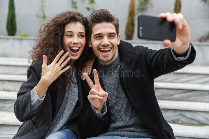 Imagem do homem dos pares e da mulher europeus 20s na roupa morna, tomando a foto do selfie no telefone celular ao sentar-se nas fotografia de stock royalty free Imagem do homem dos pares e da mulher europeus 20s na roupa morna, tomando a foto do selfie no telefone celular ao sentar-se nas fotografia de stock royalty free