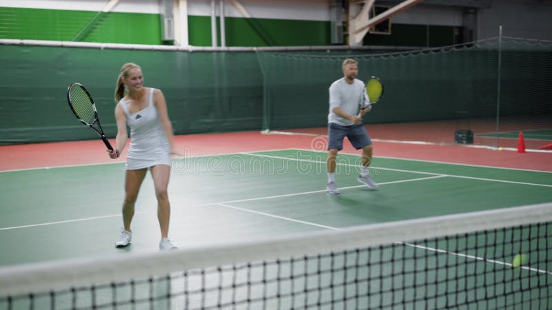 Imagem dos pares felizes que jogam o tênis na corte interna Os jovens vestidos no equipamento do esporte estão passando o tempo j
