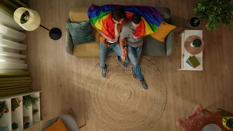 Imagen de primera plana de una pareja homosexual sentada en el sofá mirando a la cámara envuelta en una bandera de lgbt para