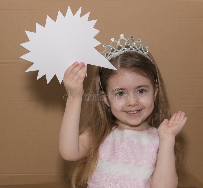 Imagen de una niña confundida con una corona aislada sobre fondo de cartón gris sosteniendo burbuja de habla foto de archivo