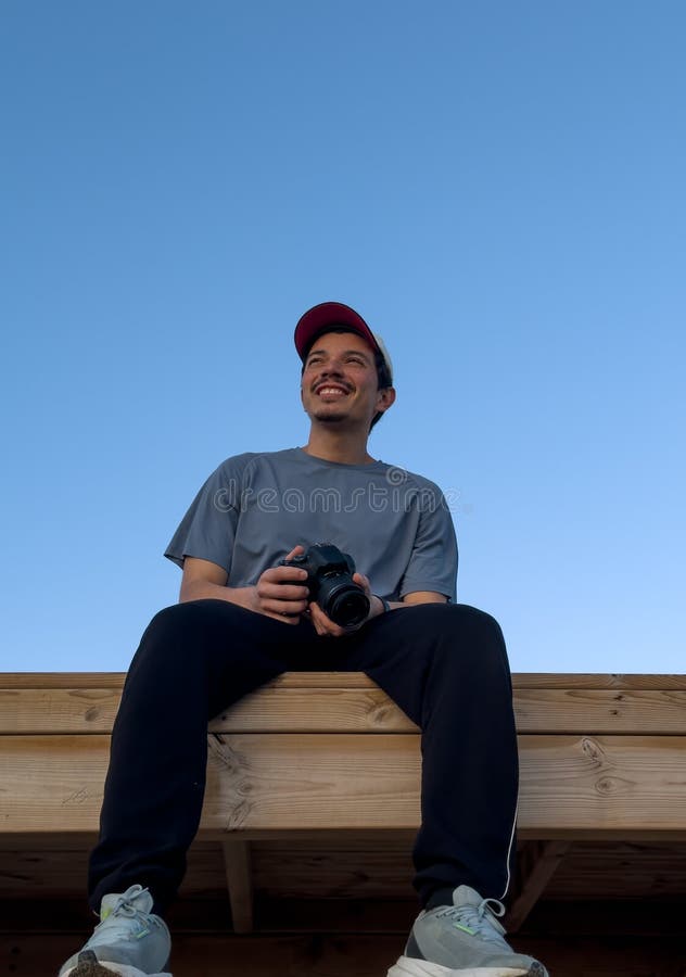Imagen vertical de un fotógrafo sonriente sentado en una estructura de madera con un cielo azul al fondo imagen de archivo