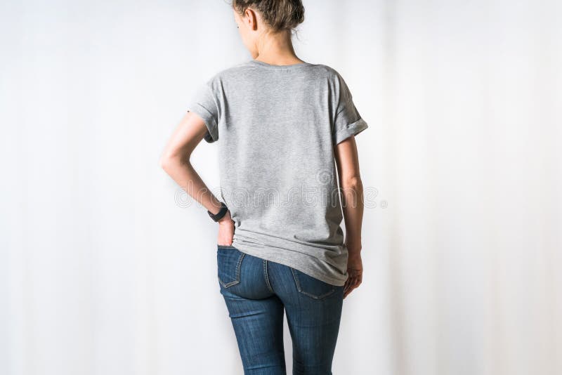 Immagine potata Retrovisione della giovane donna, vestita in maglietta grigia e blue jeans, stanti per il fondo leggero fotografia stock libera da diritti Immagine potata Retrovisione della giovane donna, vestita in maglietta grigia e blue jeans, stanti per il fondo leggero fotografia stock libera da diritti