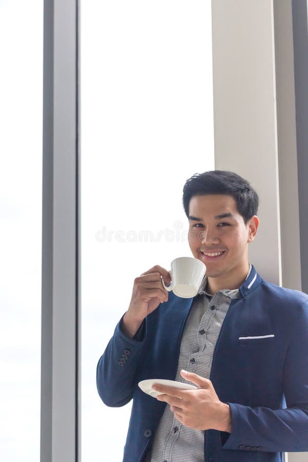 Imprenditore asiatico, in mano una tazza di caffè bianco davanti alla scrivania c'è un computer portatile fotografie stock Imprenditore asiatico, in mano una tazza di caffè bianco davanti alla scrivania c'è un computer portatile fotografie stock
