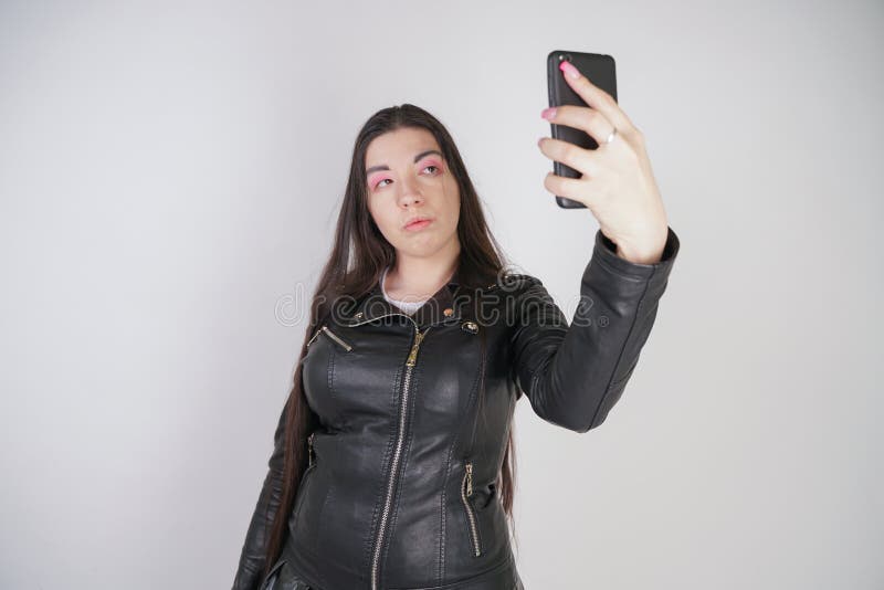 Incantare castana in bomber nero nei supporti di stile della roccia con uno smartphone su un fondo bianco nello studio immagini stock